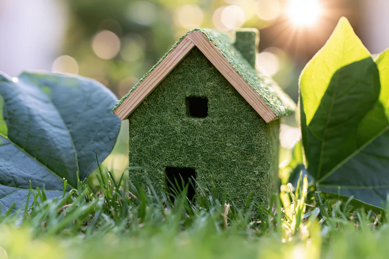 Small green textured house model sitting in fresh grass surrounded by large leaves under sunlight
