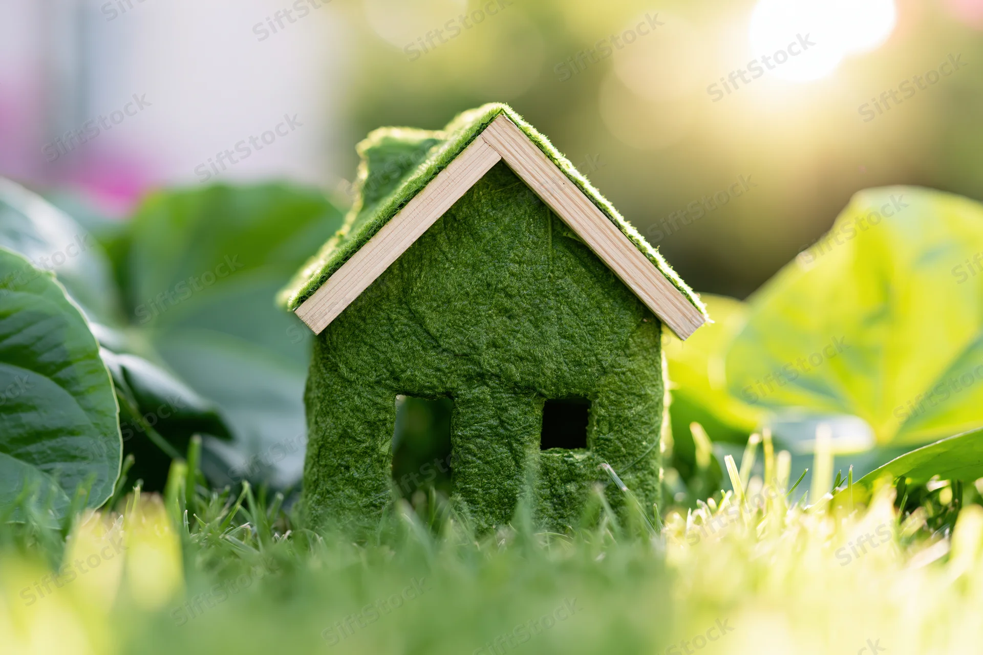 Green moss-covered house model sitting in fresh grass with blurred foliage and sunlight in background