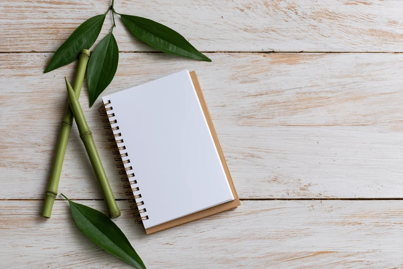 Open spiral notebook with blank white page on wooden surface beside bamboo stalks and green leaves