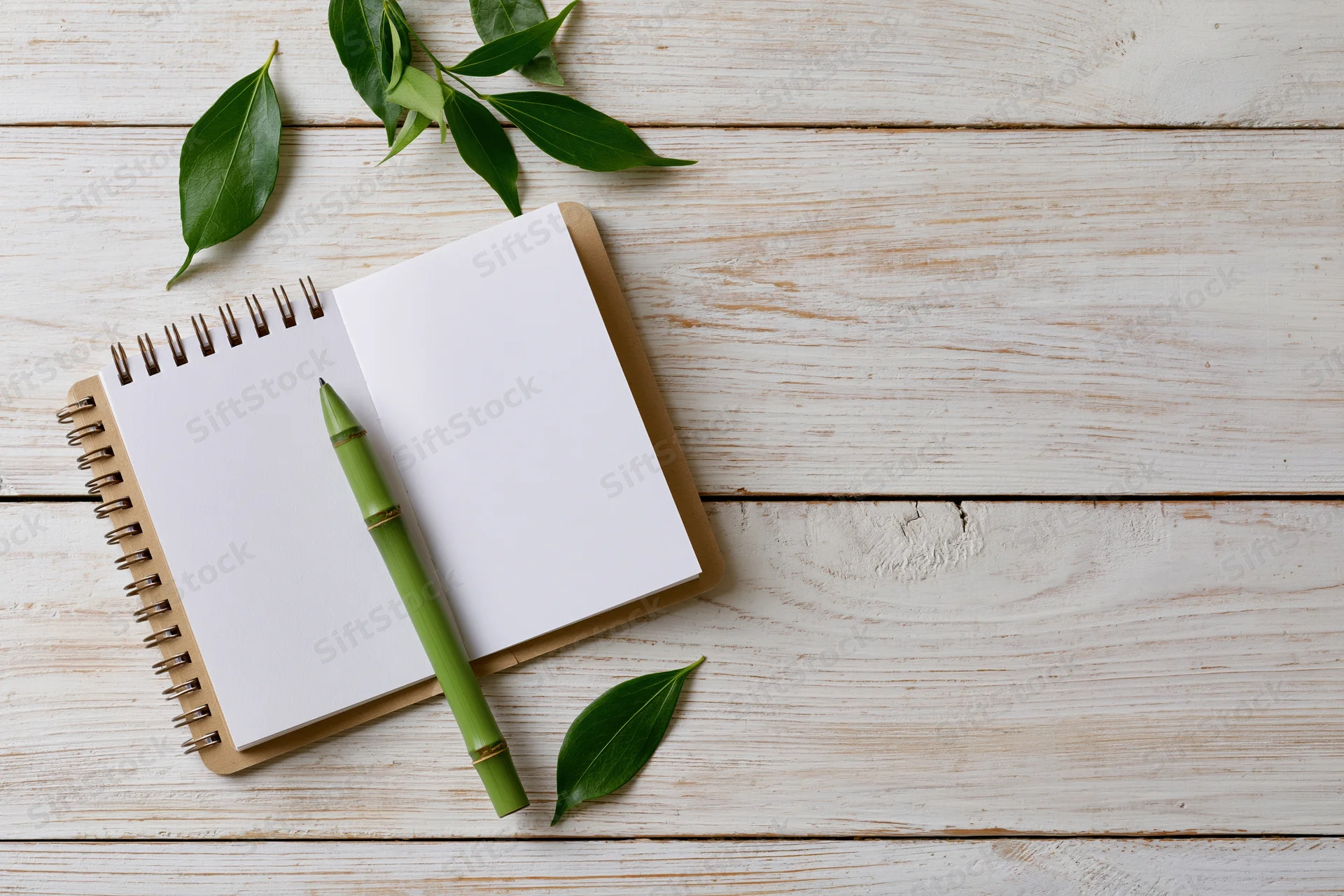 Open spiral notebook with blank white pages and green bamboo pen on wooden surface with scattered leaves