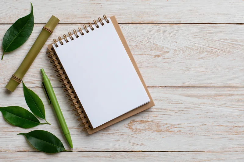 Spiral-bound kraft notebook open to a blank white page on a wooden surface with a green pen, bamboo tube, and leaves arranged beside it