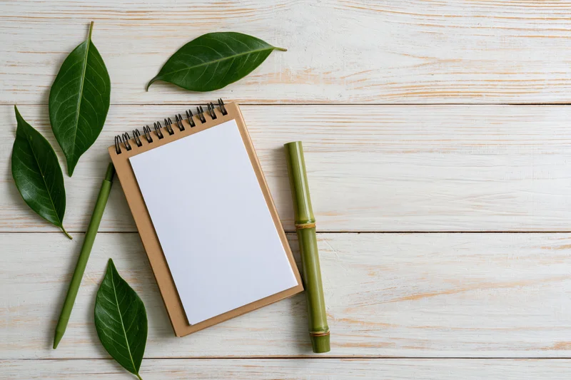 Spiral-bound kraft notebook open to a blank white page with green bamboo pen and leaves on a light wooden surface
