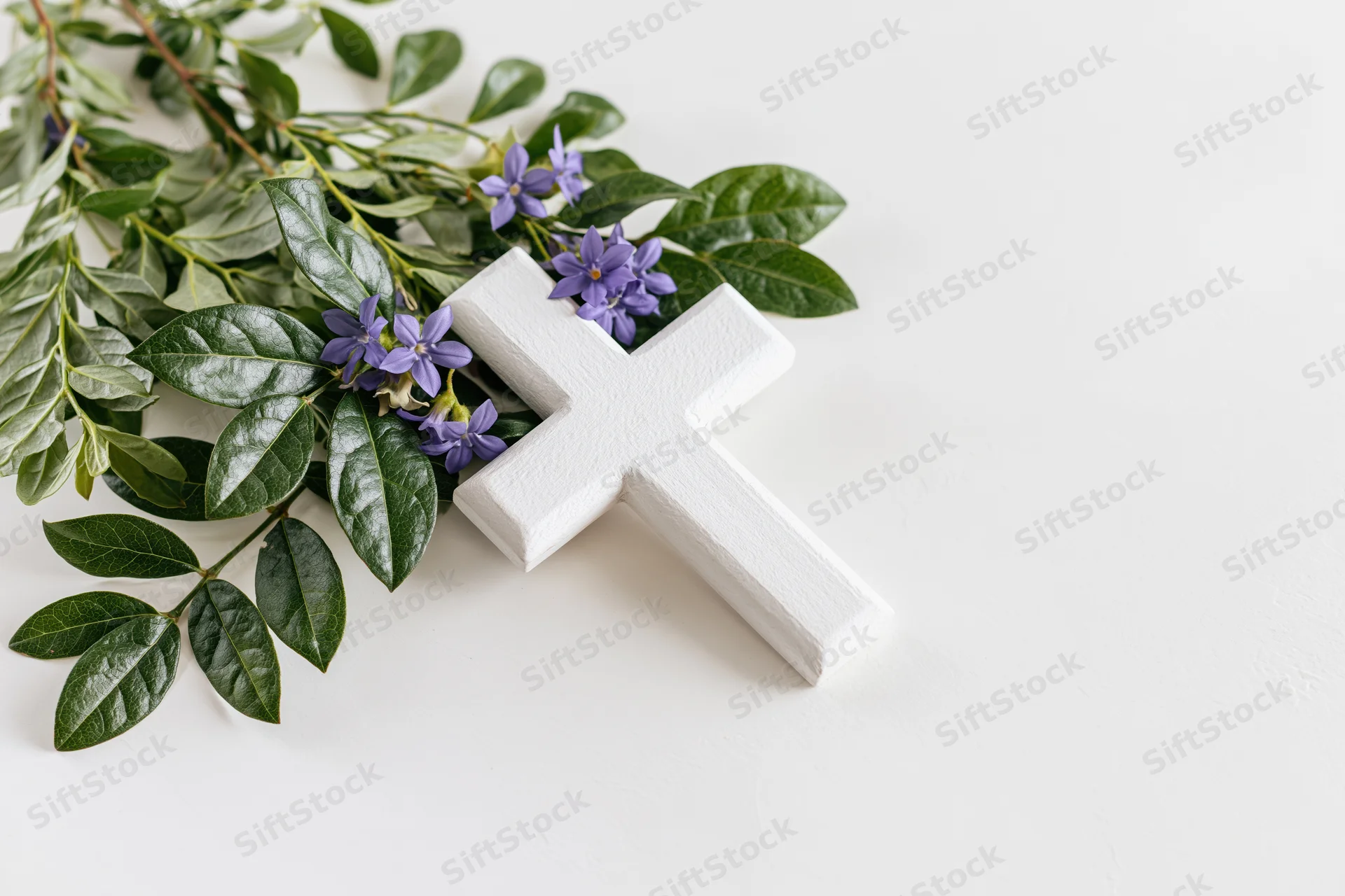 Small white wooden christian cross lying flat on white surface with green leaves and purple flowers arranged beside it