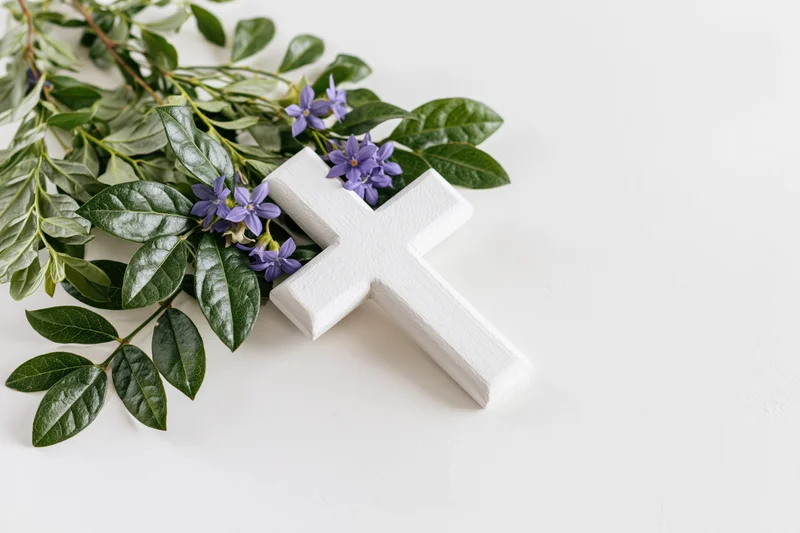 Small white wooden christian cross lying flat on white surface with green leaves and purple flowers arranged beside it