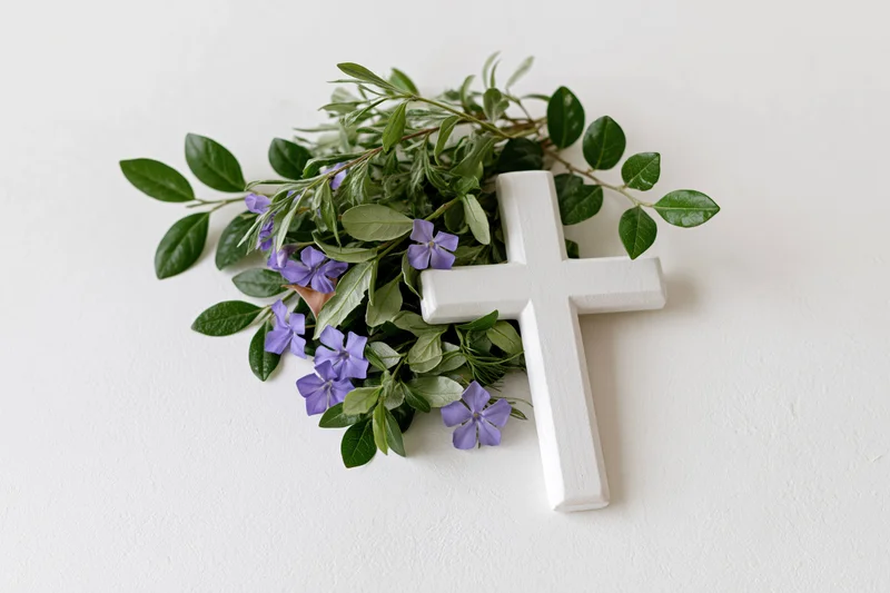 Small white wooden christian cross lying flat on white surface with purple flowers and green foliage arranged beside it