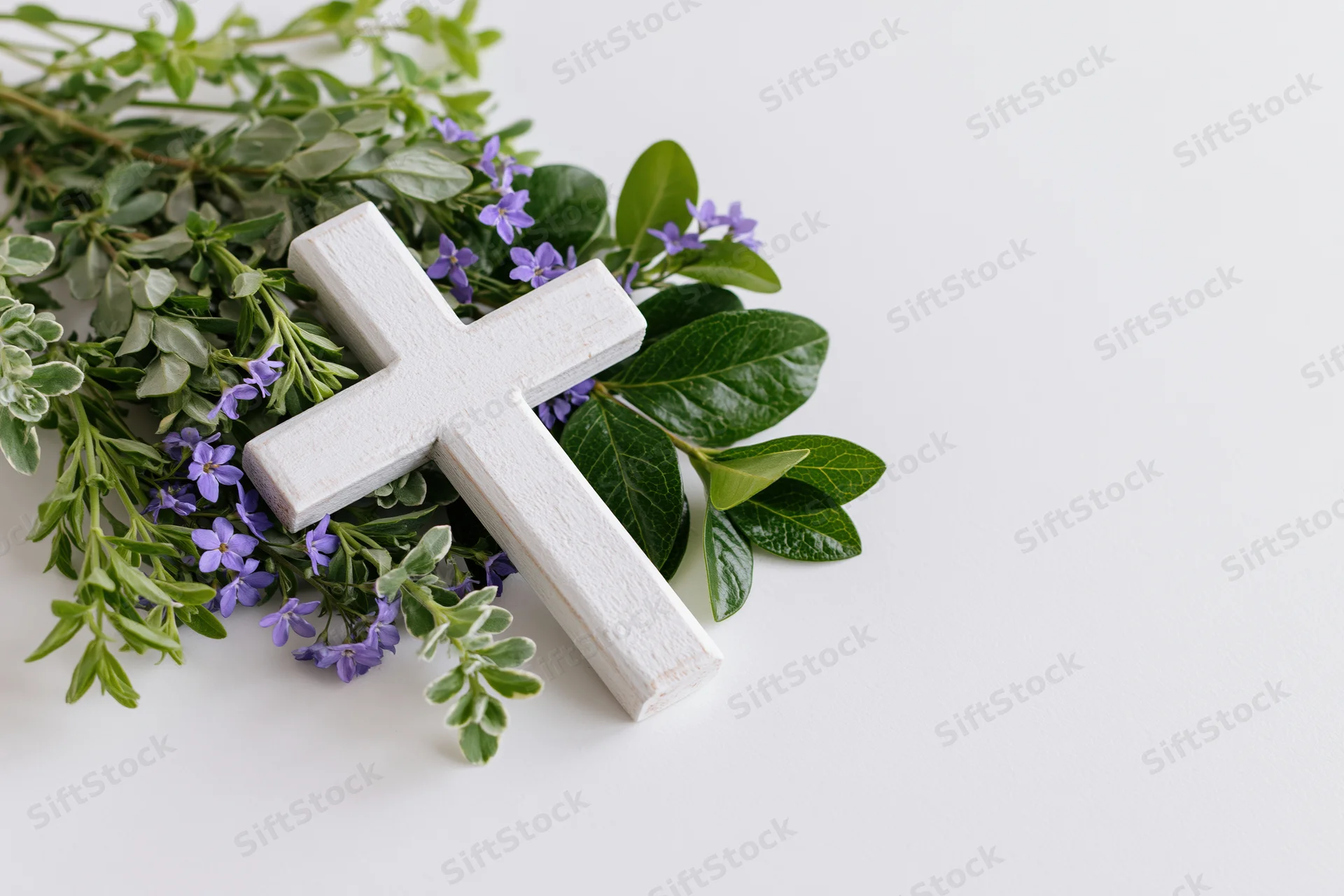 Small white wooden christian cross lying flat on white surface surrounded by green leaves and purple flowers