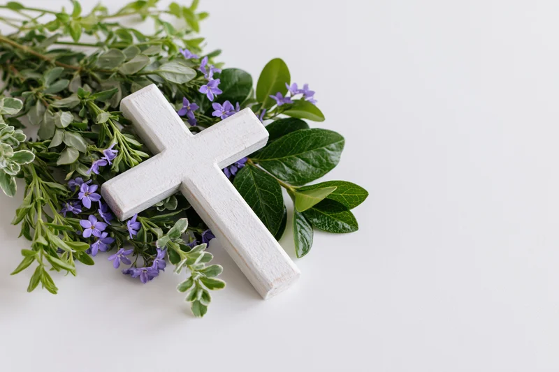 Small white wooden christian cross lying flat on white surface surrounded by green leaves and purple flowers