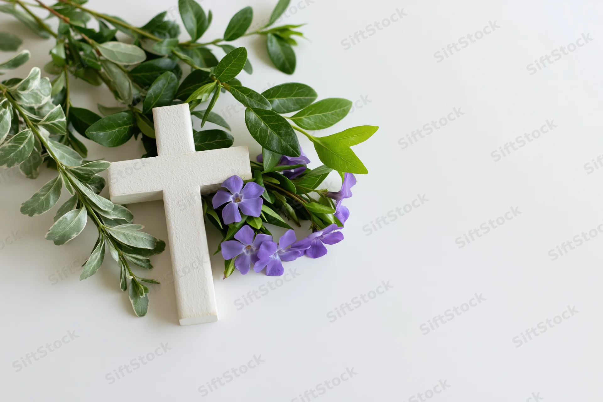 White wooden christian cross lying flat on white surface with purple flowers and green foliage arranged beside it