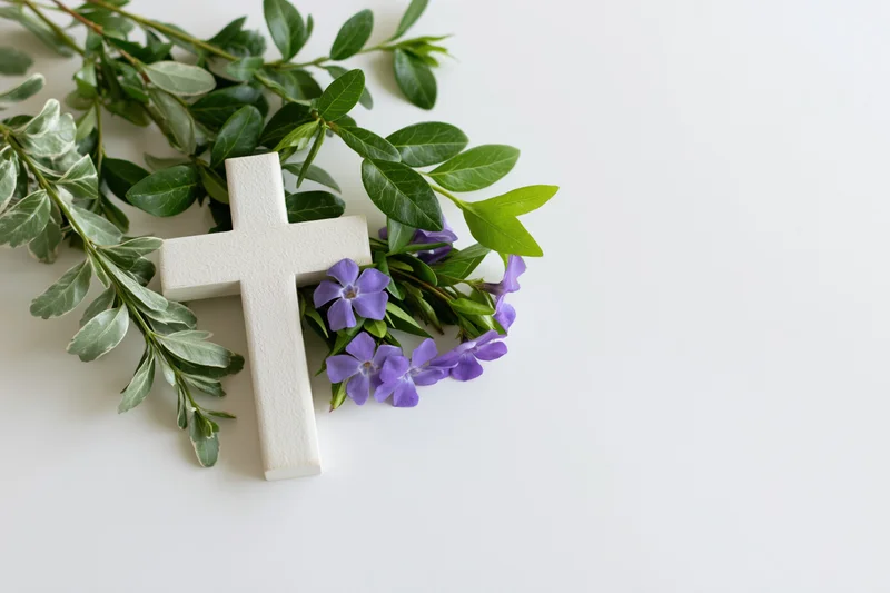 White wooden christian cross lying flat on white surface with purple flowers and green foliage arranged beside it