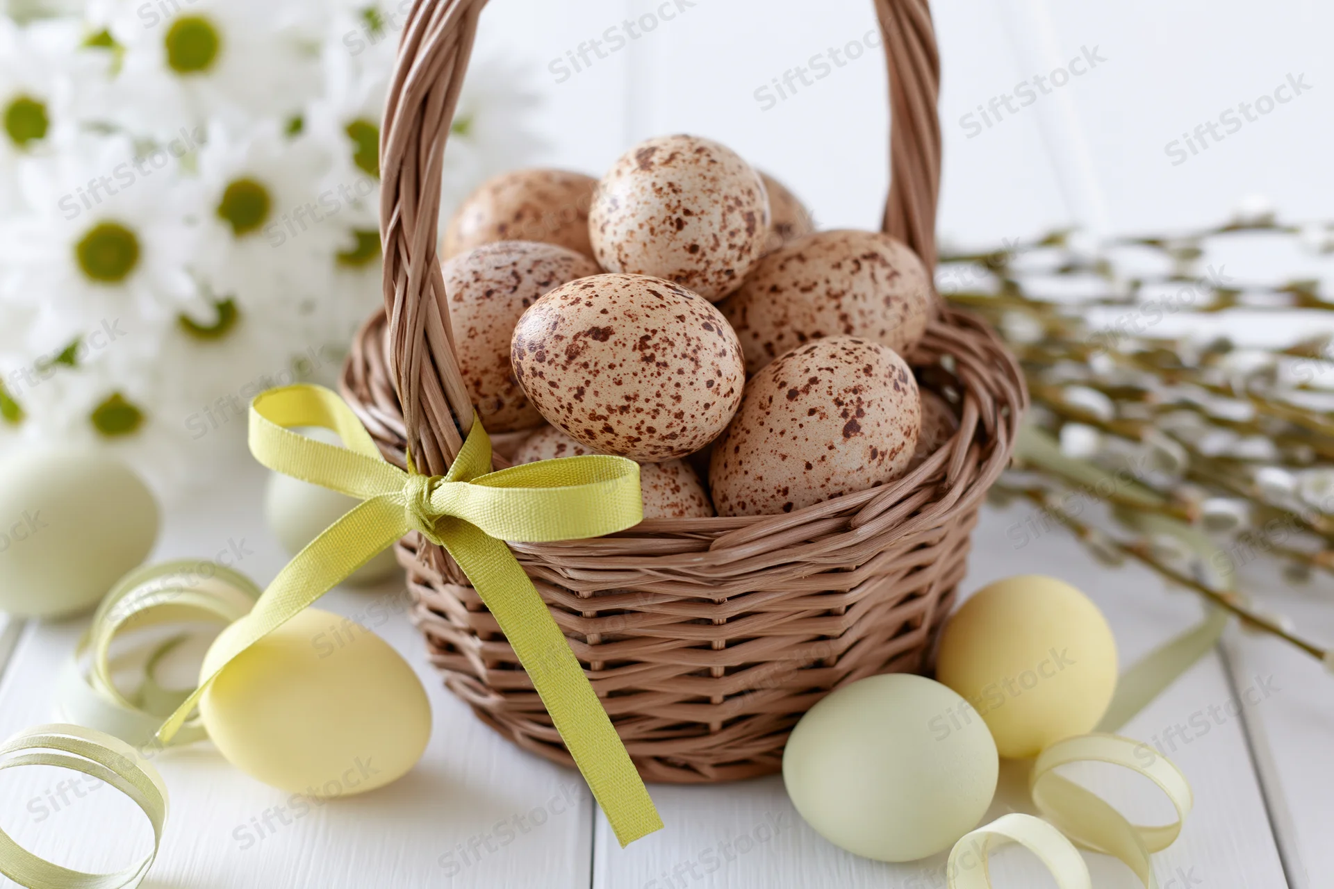 Wicker basket filled with speckled natural eggs tied with yellow ribbon on white table with spring flowers and pastel eggs around