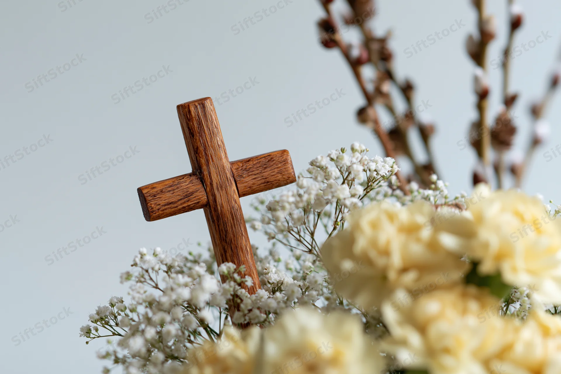 Wooden cross with visible grain and warm honey tones nestled among white baby's breath and cream roses in a floral arrangement