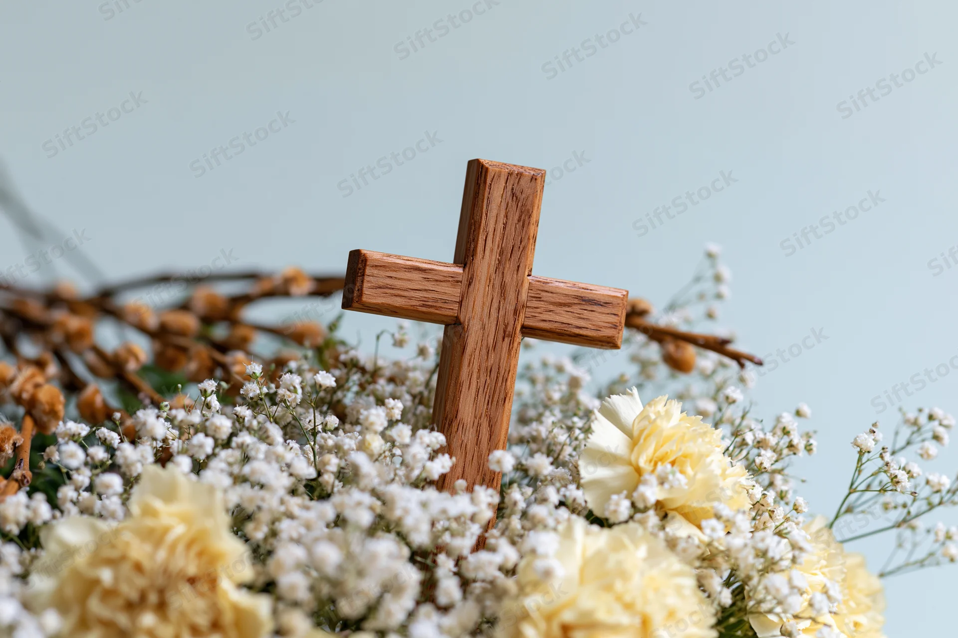 Wooden cross with visible grain and warm honey tones nestled in a bouquet of white baby's breath and yellow carnations