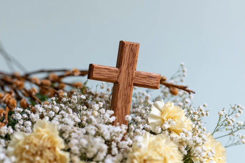 Wooden cross with visible grain and warm honey tones nestled in a bouquet of white baby's breath and yellow carnations