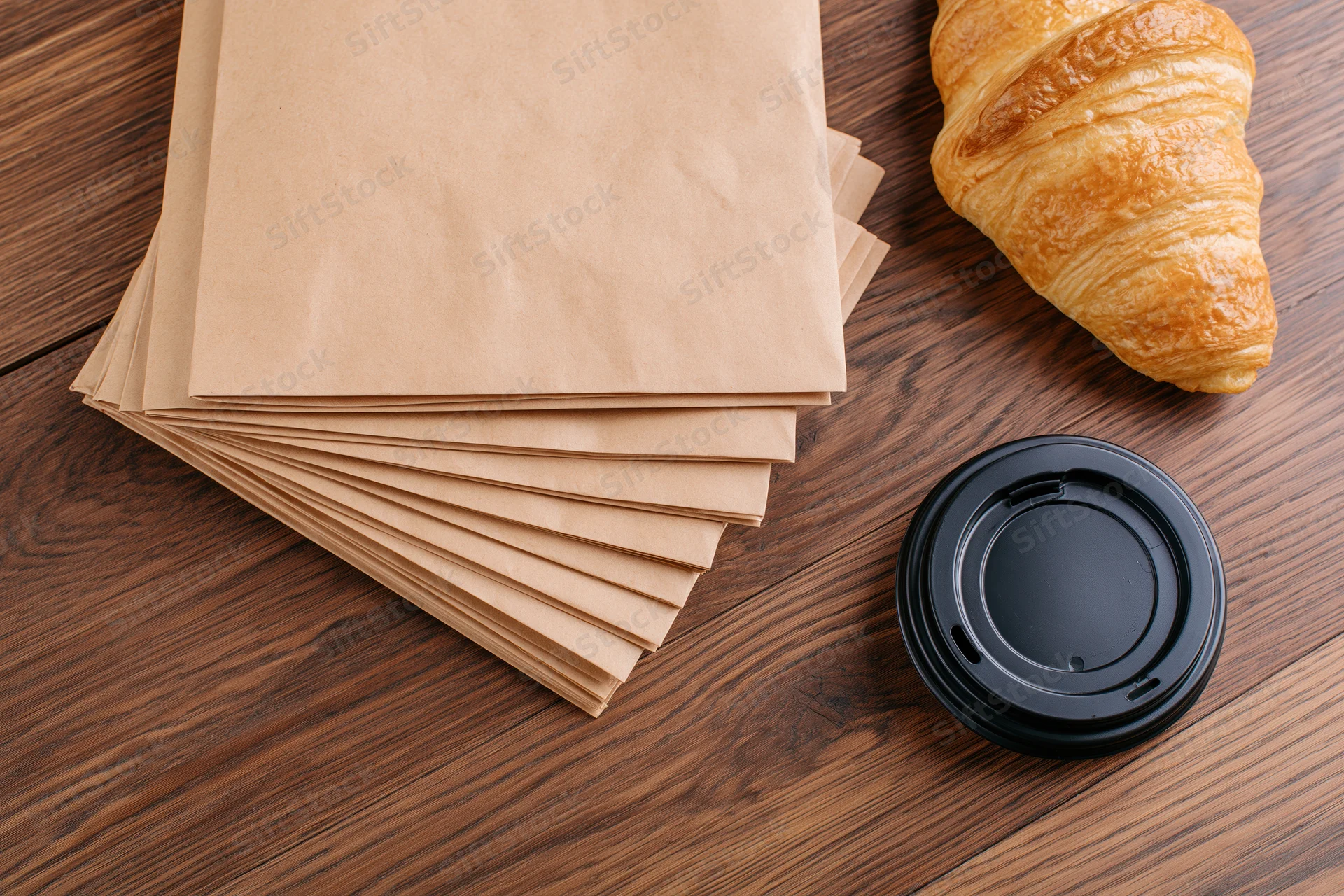 Stack of blank kraft paper bags fanned out on wooden table with croissant and coffee cup lid nearby