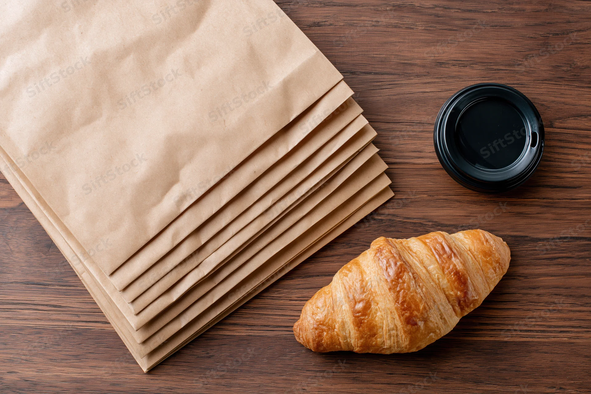 Stack of kraft paper bags fanned out on wooden table with croissant and coffee cup beside them