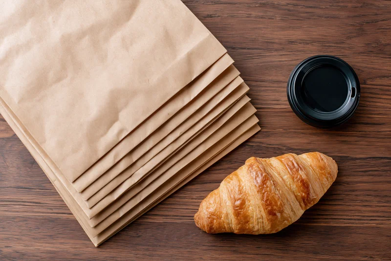 Stack of kraft paper bags fanned out on wooden table with croissant and coffee cup beside them