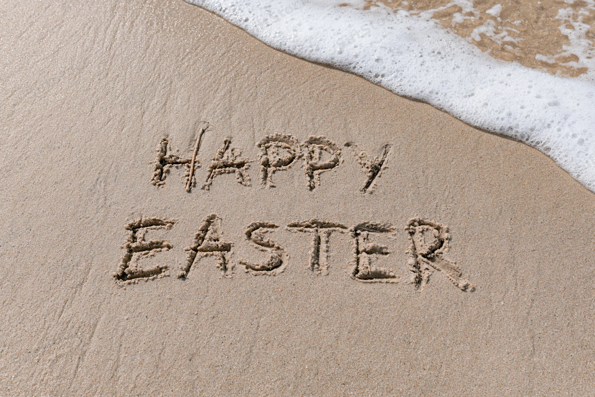 Happy easter written in wet sand on a beach with ocean foam approaching