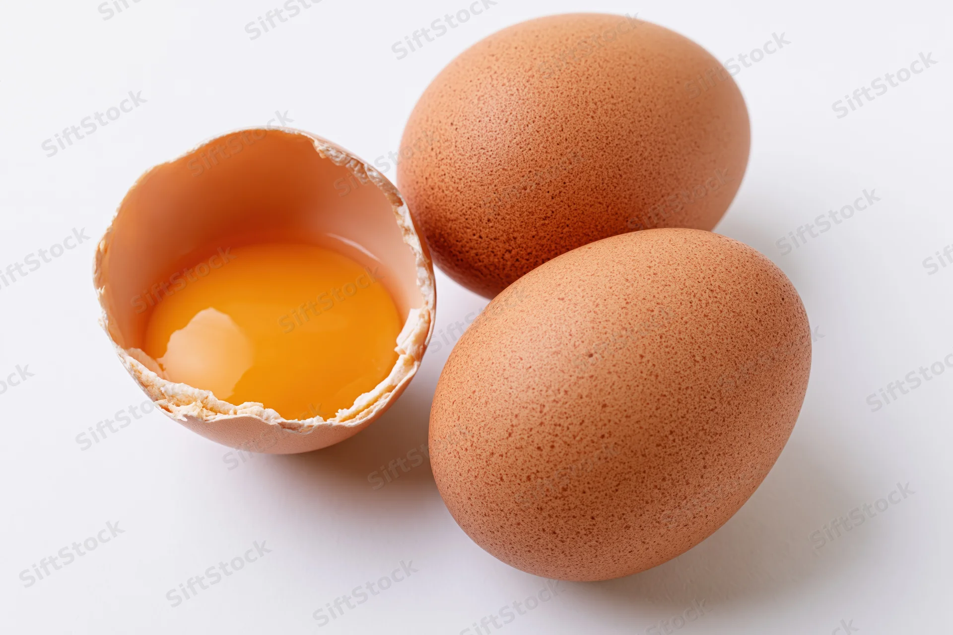 Three brown chicken eggs with one cracked open showing yolk on white background