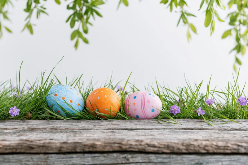 Three painted easter eggs nestled in green grass on a wooden surface with purple flowers and hanging leaves above