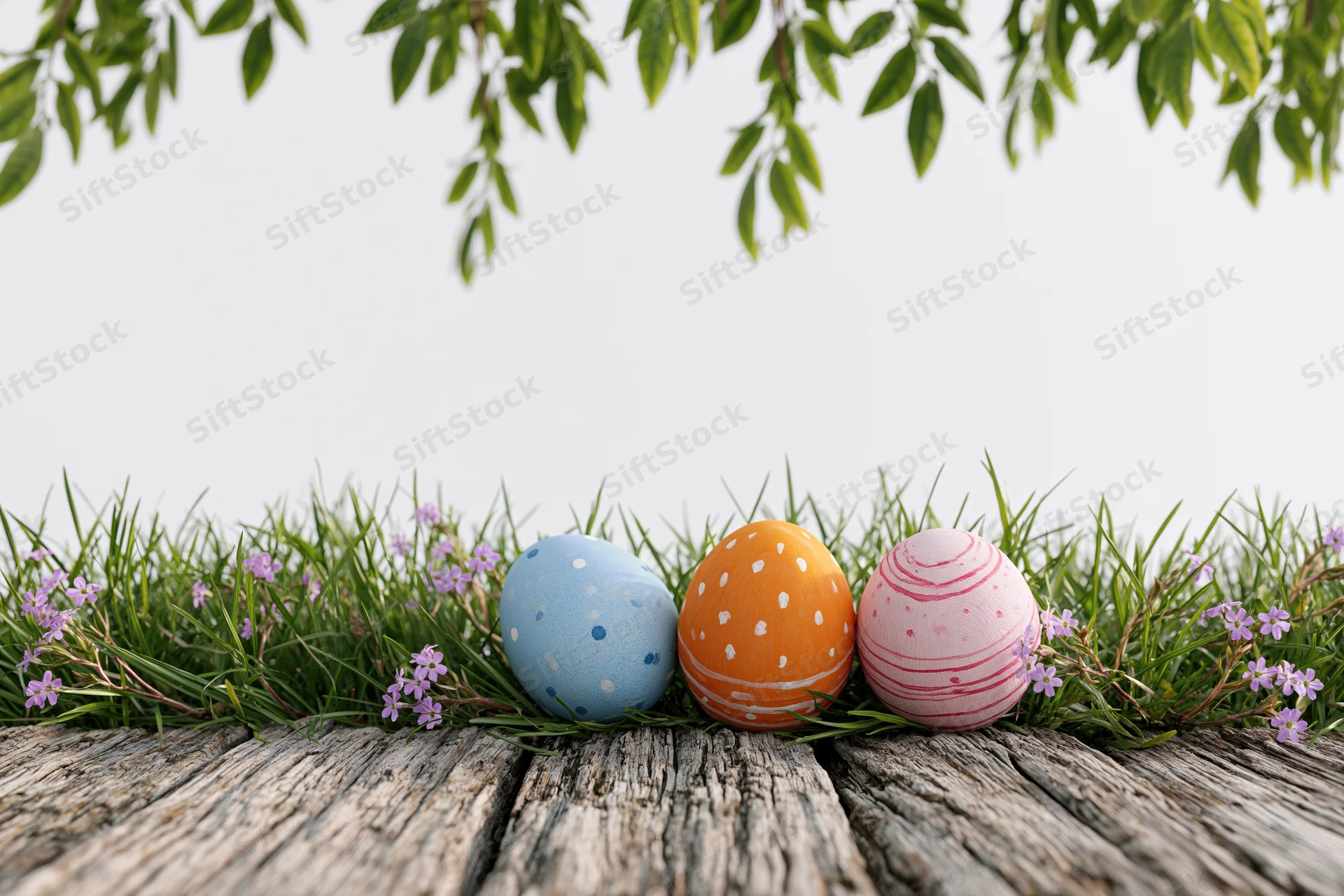 Three painted easter eggs nestled in green grass with purple flowers on a wooden surface