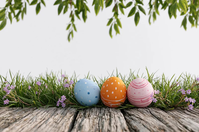 Three painted easter eggs nestled in green grass with purple flowers on a wooden surface