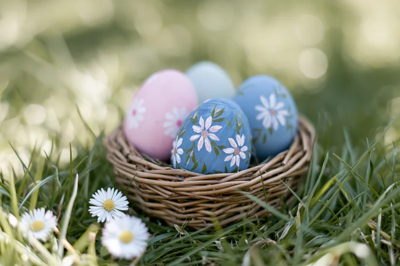 Pastel-painted easter eggs with floral designs in a wicker basket on grass with daisies nearby