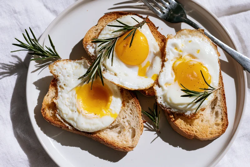 Three sunny-side-up eggs on toasted bread slices arranged on a white plate with rosemary sprigs and a fork