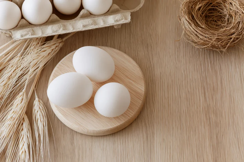 Three white eggs arranged on a small round wooden plate on a light wooden table with wheat stalks and a straw nest nearby