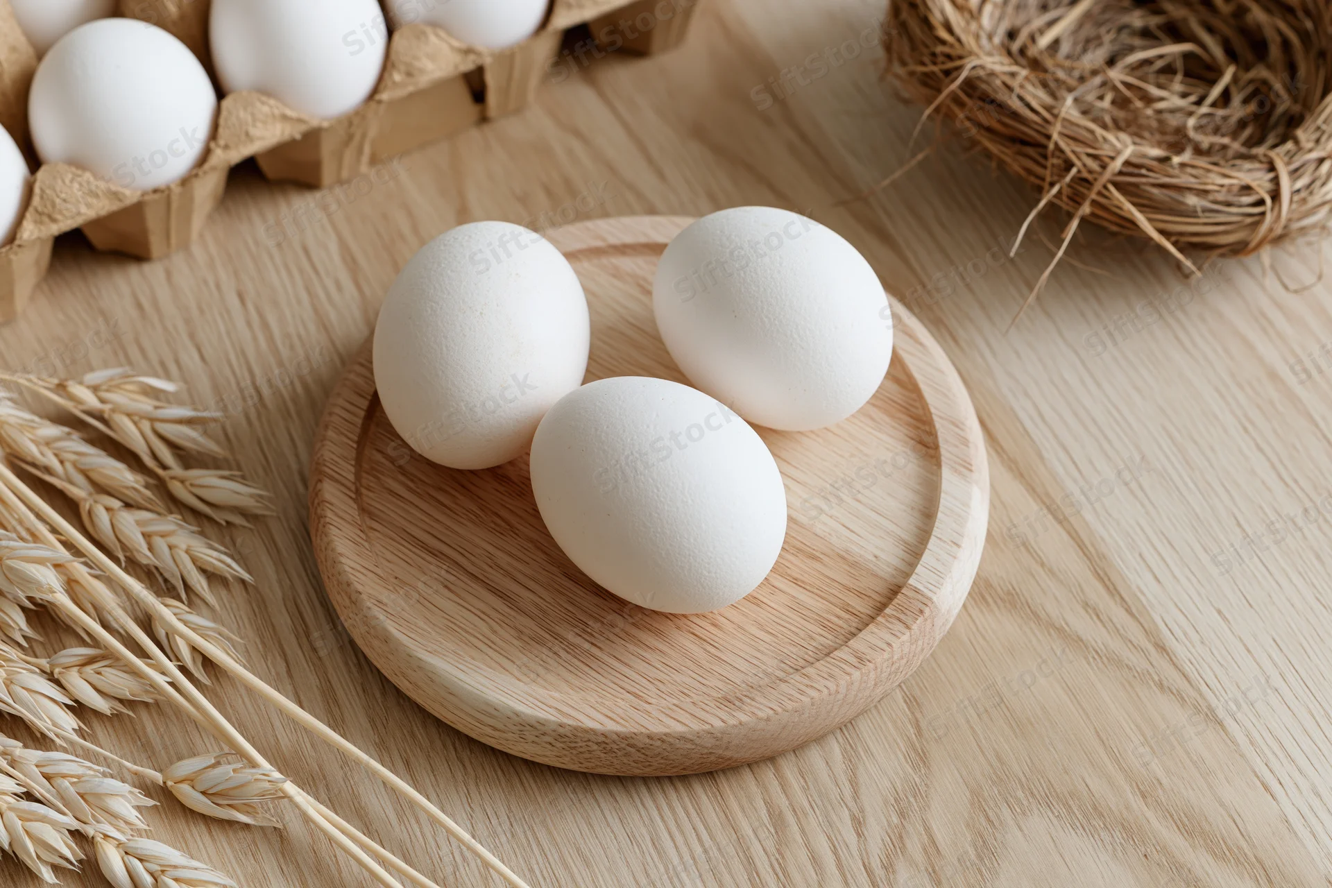 Three white eggs arranged on a small round wooden plate on a light wooden table with wheat stalks and a straw nest nearby