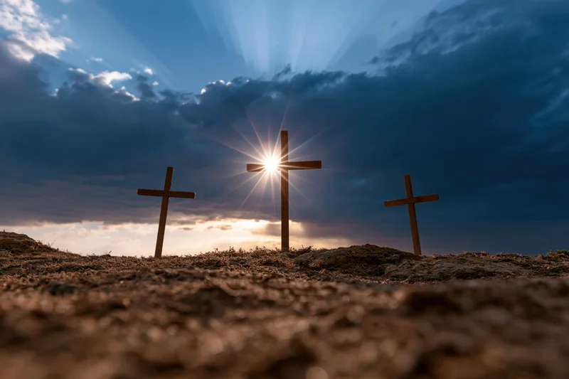 Three wooden crosses standing on a barren rocky hill under a dramatic sky with sun rays breaking through clouds