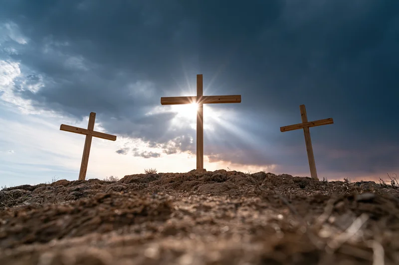 Three wooden crosses standing on a barren rocky hill under a dramatic sky with sun rays breaking through clouds
