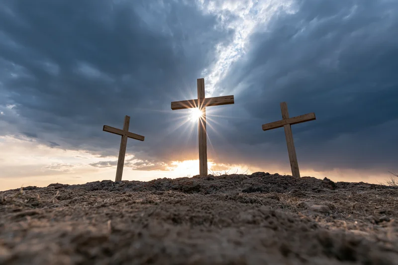Three wooden crosses standing on a barren rocky hill under dramatic sky with sun rays breaking through clouds