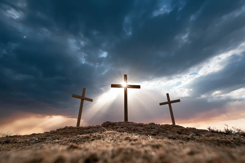 Three wooden crosses standing on a barren rocky hill under dramatic sky with sun rays breaking through clouds