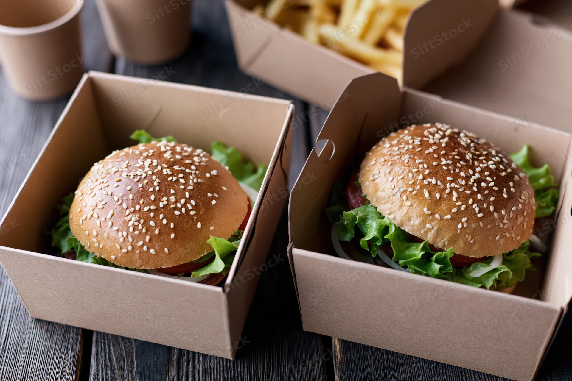 Two gourmet burgers with sesame seed brioche buns and lettuce in cardboard boxes on a dark wooden table