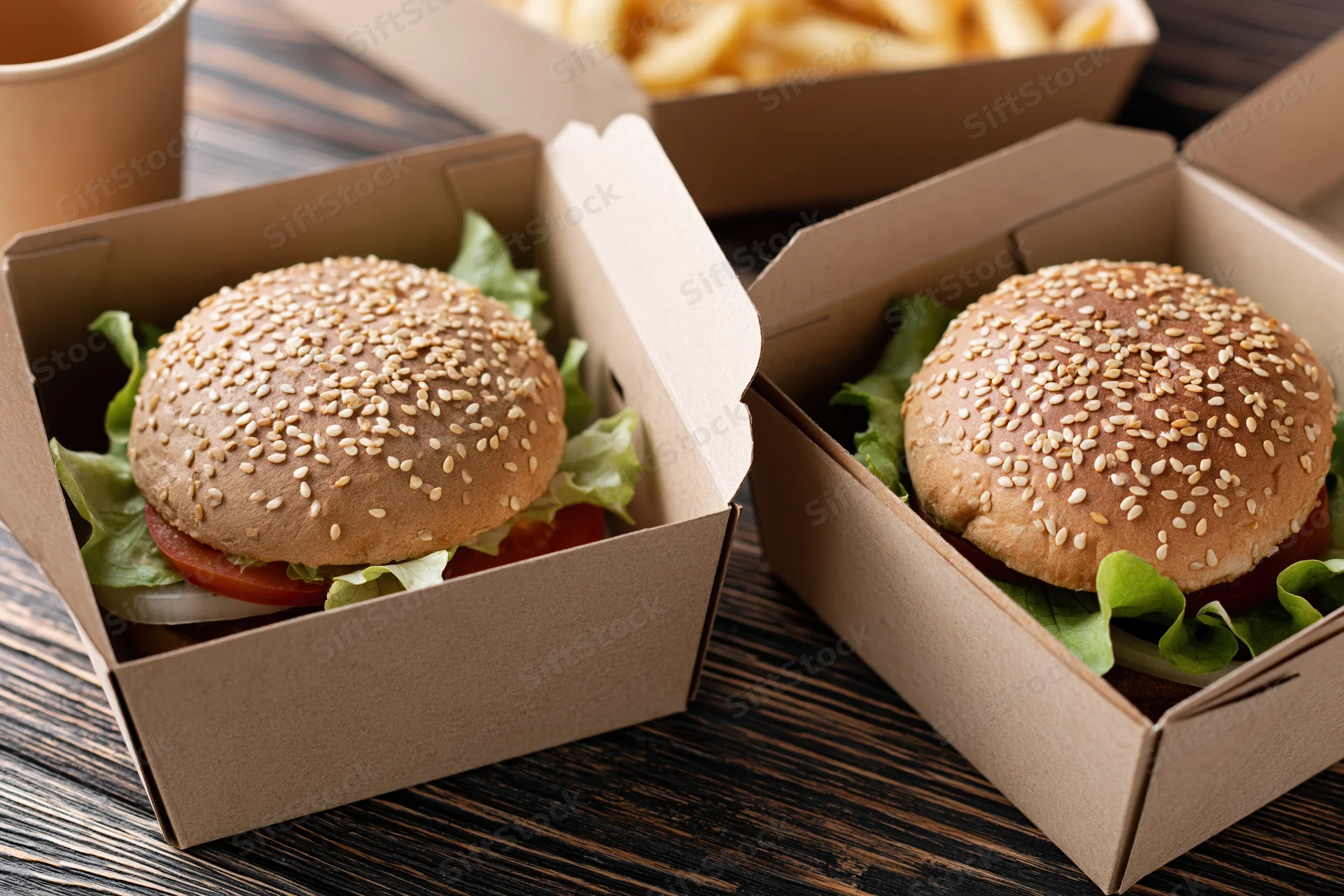 Two gourmet burgers with sesame seed brioche buns layered with lettuce, tomato, onion, and cheese in open cardboard boxes on a dark wooden table
