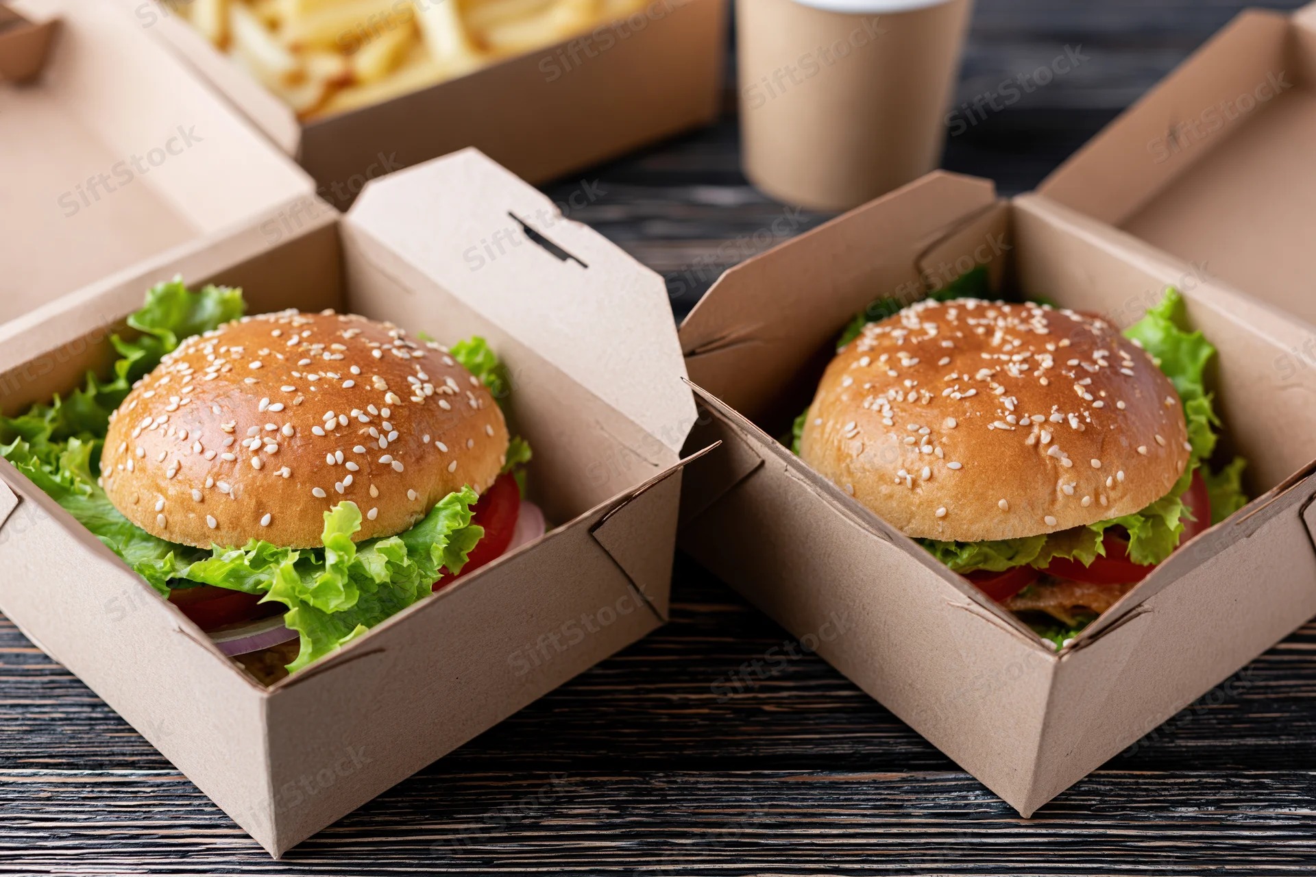 Two gourmet burgers with sesame seed brioche buns layered with lettuce, tomato, and onion in open cardboard boxes on a dark wooden table