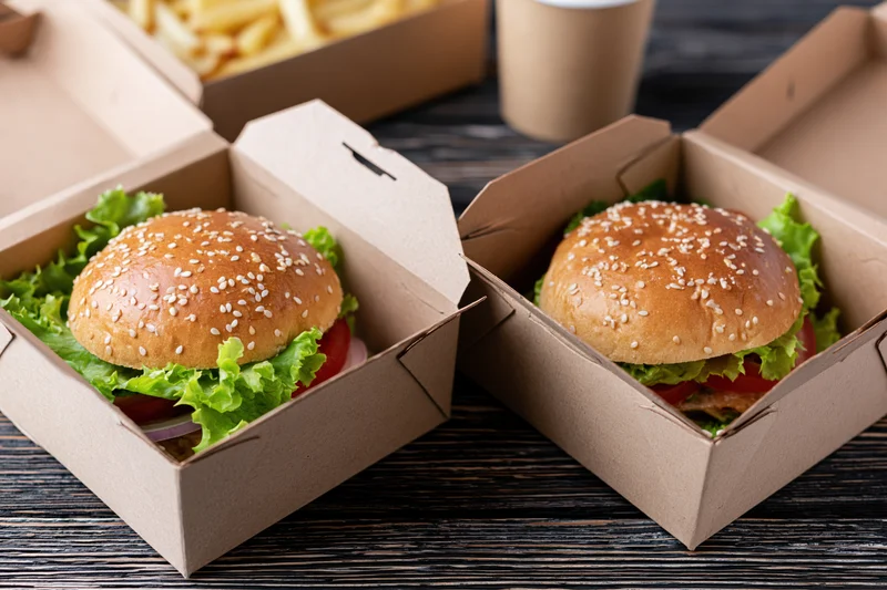 Two gourmet burgers with sesame seed brioche buns layered with lettuce, tomato, and onion in open cardboard boxes on a dark wooden table