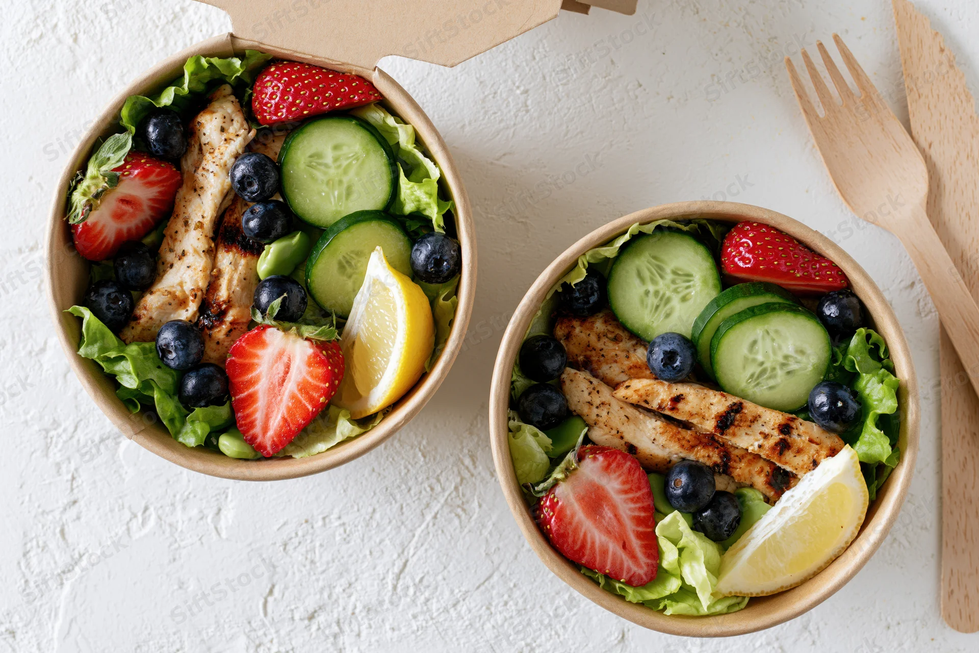 Two kraft paper takeout containers filled with fresh salads arranged on a white surface with wooden utensils beside them