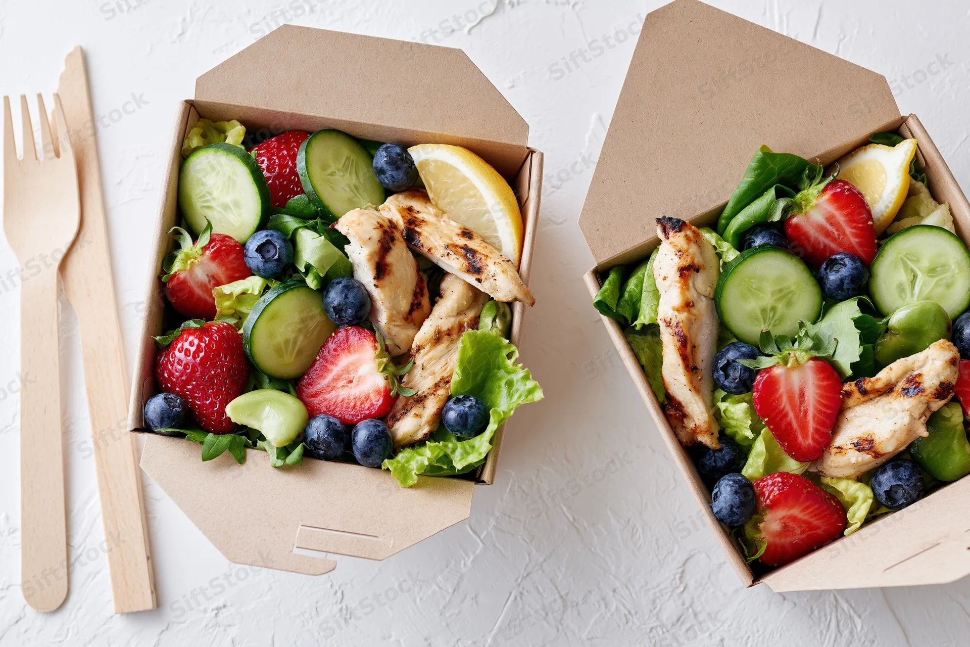 Two kraft paper takeout containers filled with fresh salads topped with grilled chicken, strawberries, blueberries, cucumbers, and lemon slices on a white surface with wooden utensils beside them