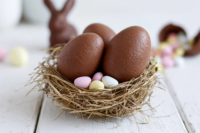 Two large textured milk chocolate easter eggs nestled in a straw nest with pastel candies on a white wooden table