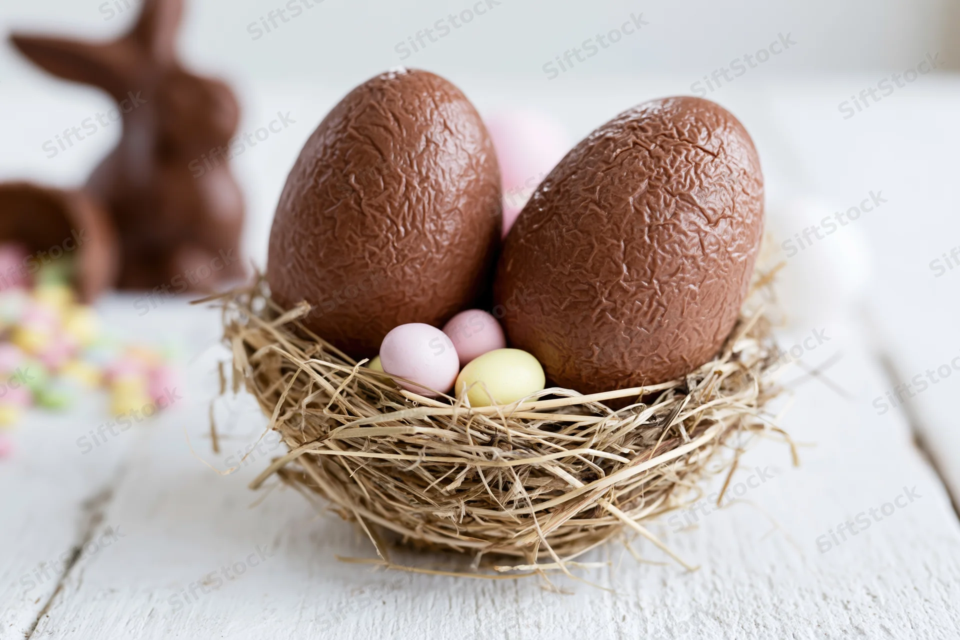 Two large textured milk chocolate easter eggs nestled in a hay nest on a white wooden table with small pastel eggs and a chocolate bunny in the background