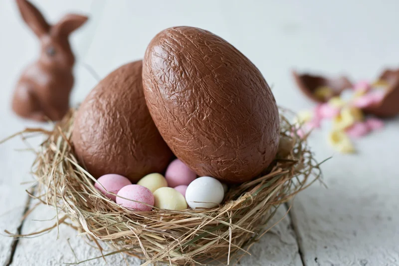 Two large textured milk chocolate easter eggs nestled in a hay nest with small pastel eggs and a chocolate bunny nearby