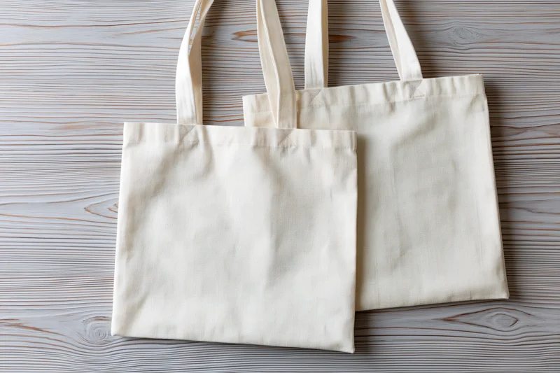 Two plain cream canvas tote bags laid flat and slightly overlapping on a wooden surface