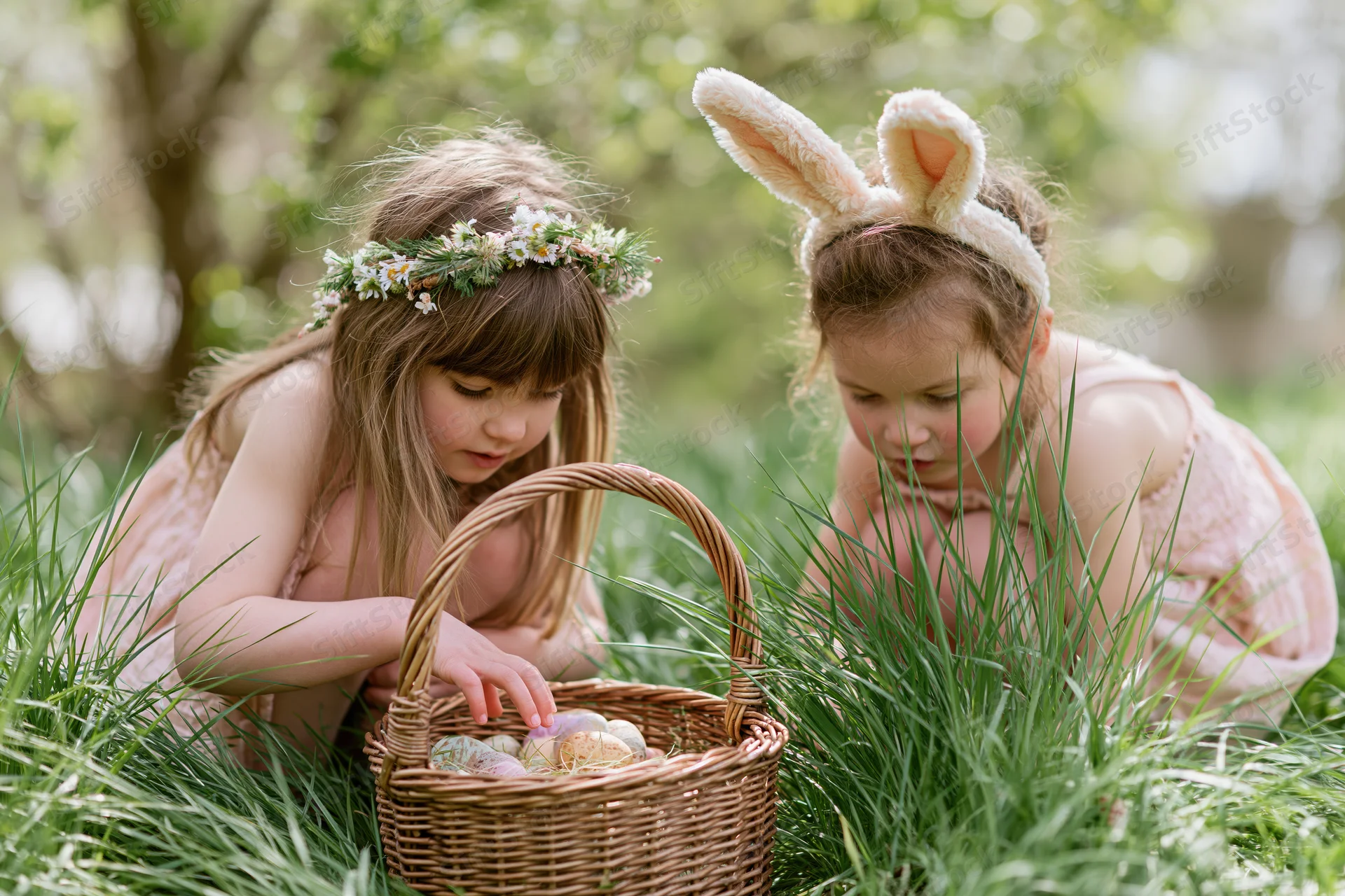 Two young girls crouching in tall grass examining a wicker basket filled with decorated eggs during an easter egg hunt