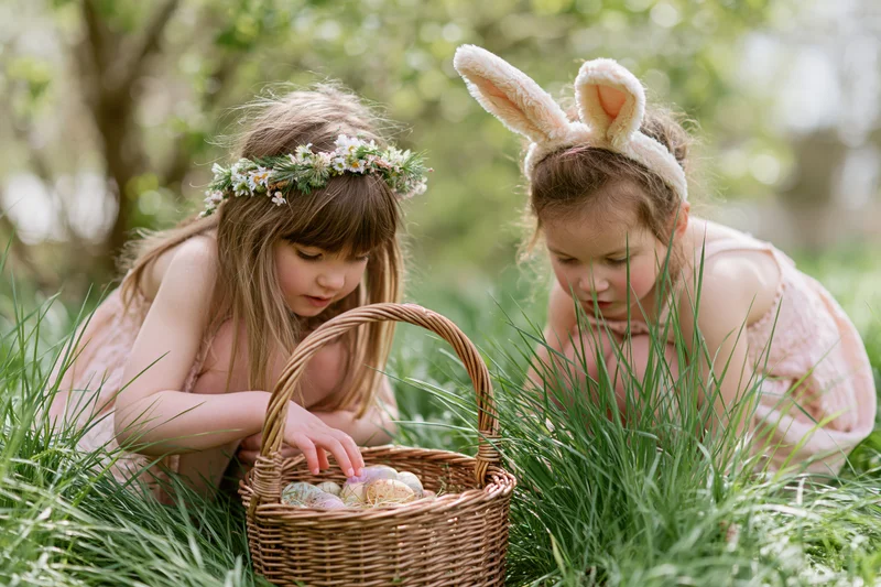 Two young girls crouching in tall grass examining a wicker basket filled with decorated eggs during an easter egg hunt
