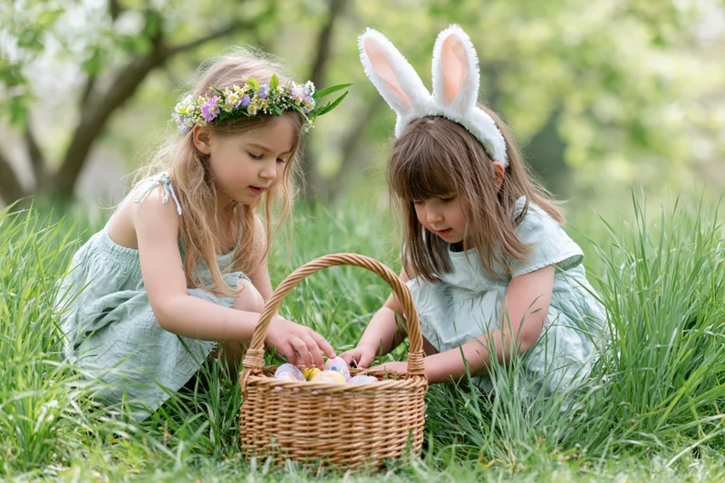 Two young girls crouching in tall grass examining a wicker basket filled with decorated eggs during an easter celebration