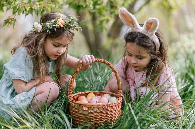 Two young girls crouching in tall grass examining a wicker basket filled with colorful easter eggs