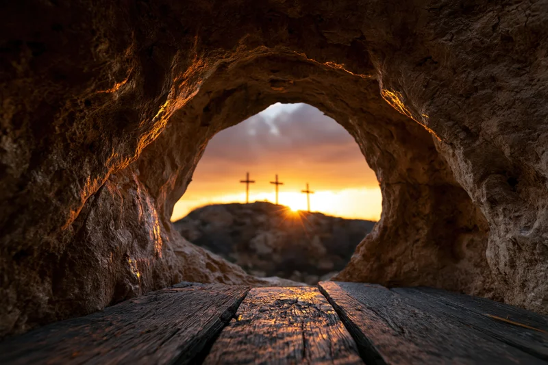 View from inside a stone cave looking out at three crosses on a hill at sunset through a rounded opening with wooden planks on the floor