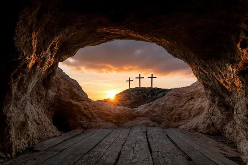 View from inside a dark stone cave looking out at three crosses on a hill at sunset through a rounded opening with wooden floor and rocky walls