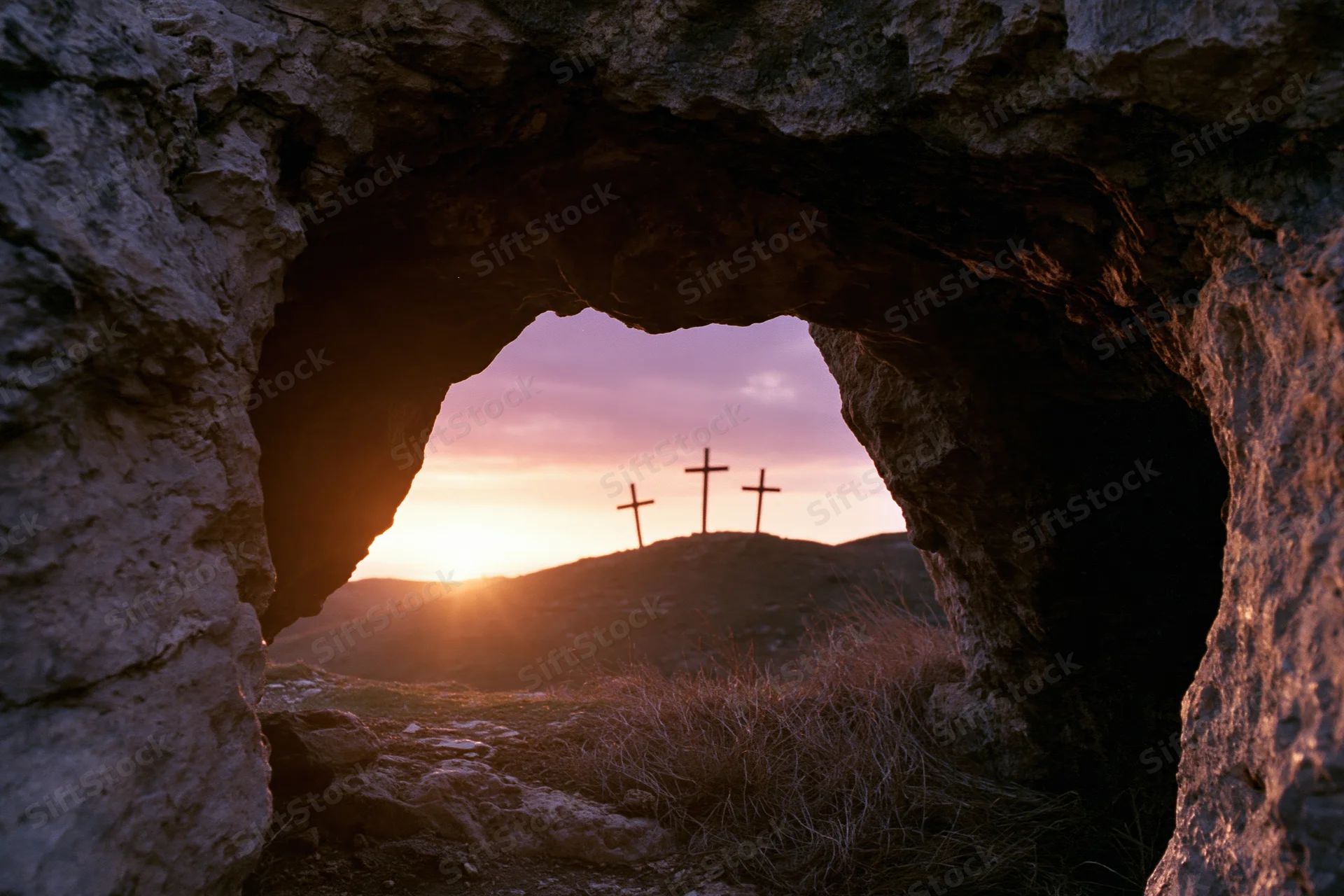 View from inside a dark stone cave looking outward through a natural arch at three crosses silhouetted on a hill at sunset